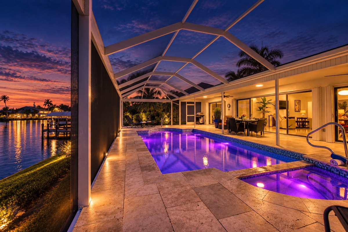 Purple-lit screened pool beside waterfront home at sunset